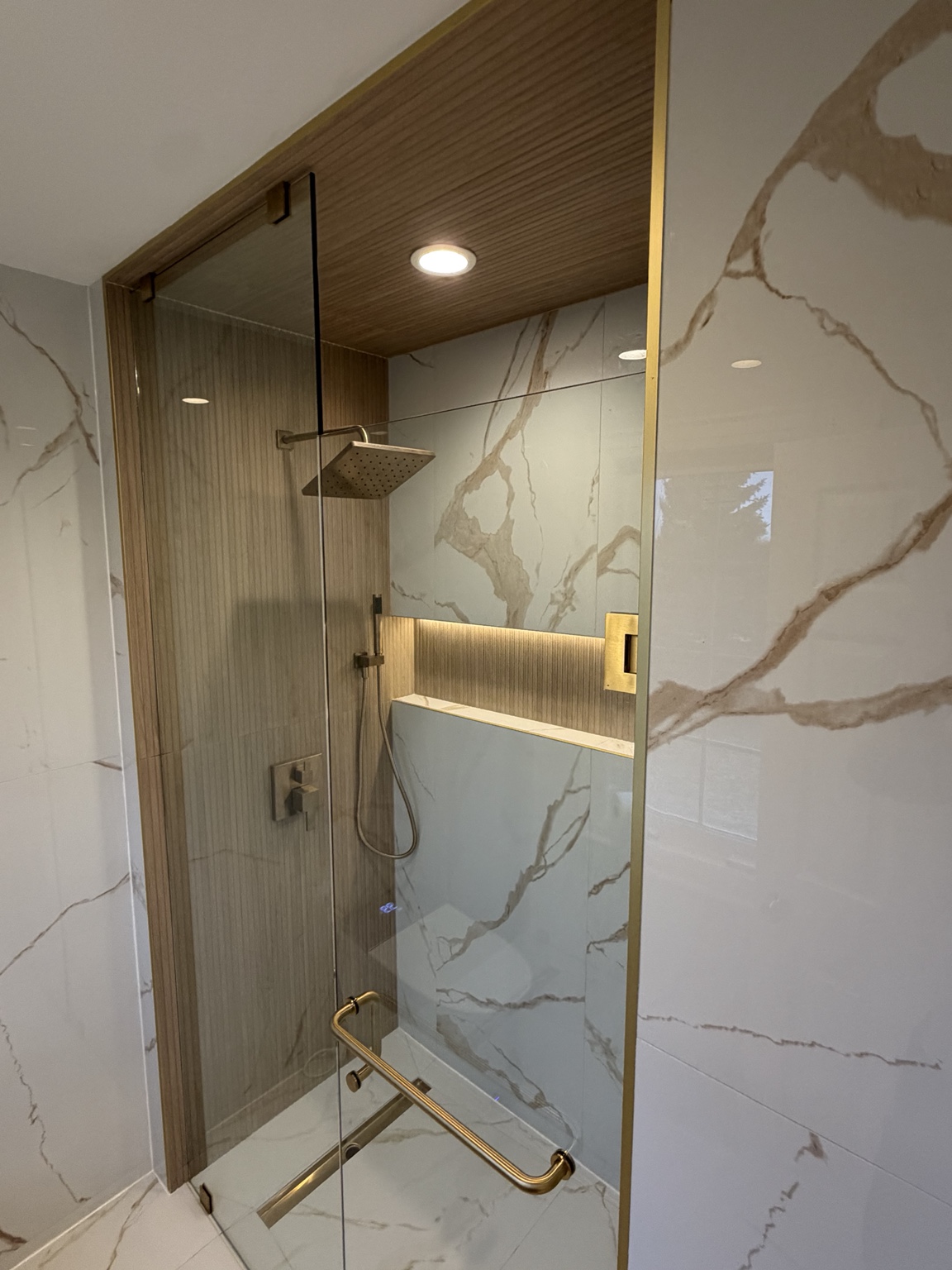 Glass shower with gold fixtures, marble-look tiles and wood-look ceiling